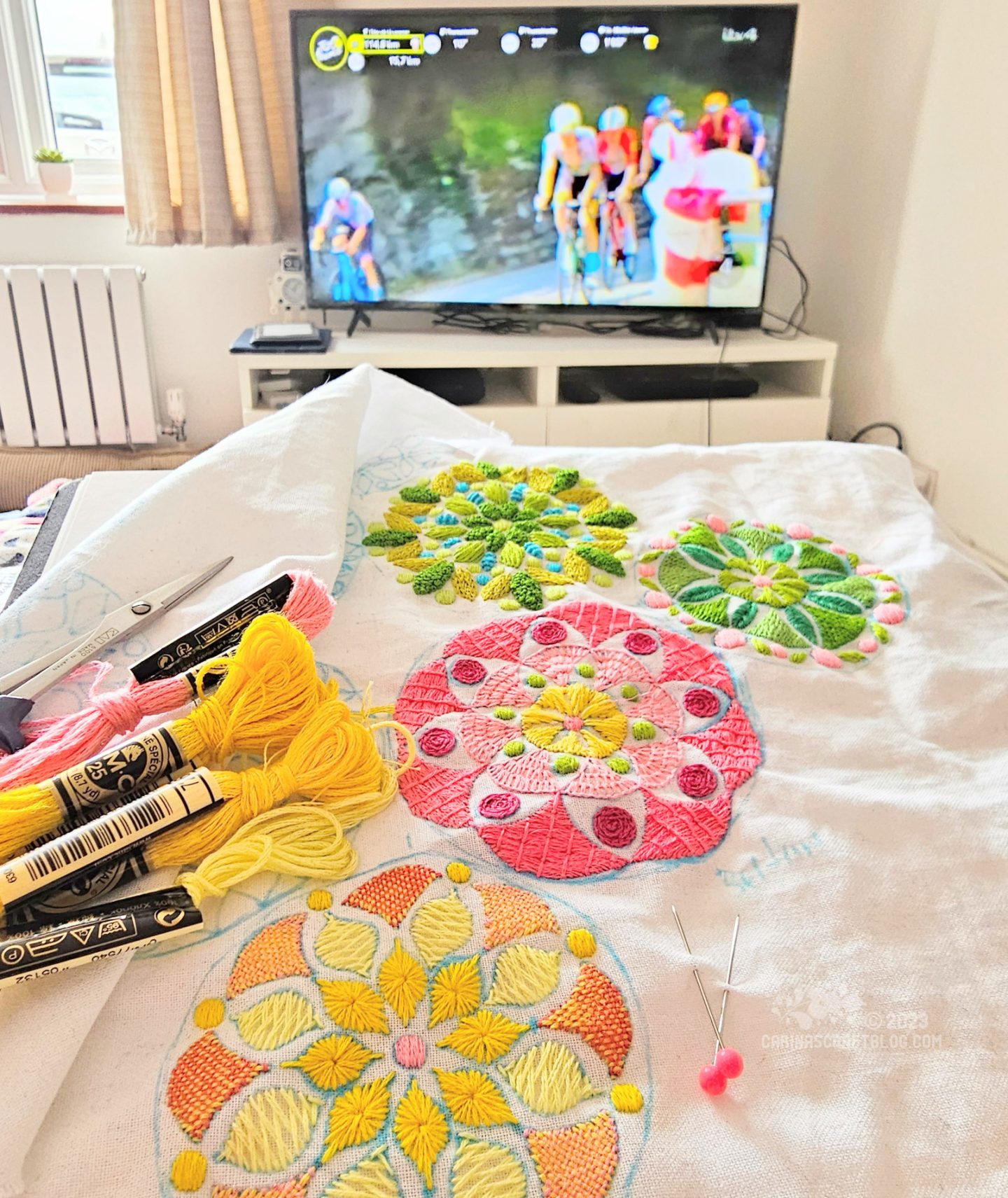 Close view of an embroidery on white fabric with mandala inspired motifs in yellow, pink and green colours. In the background is a tv showing the Tour de France bike race.