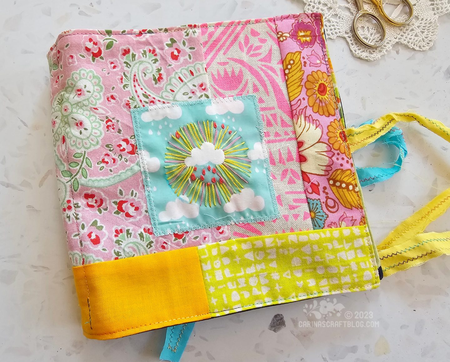 Overhead view of the front cover of a textile book. It is made of randomly sized squares and rectangles of pink, yellow and aqua fabrics.