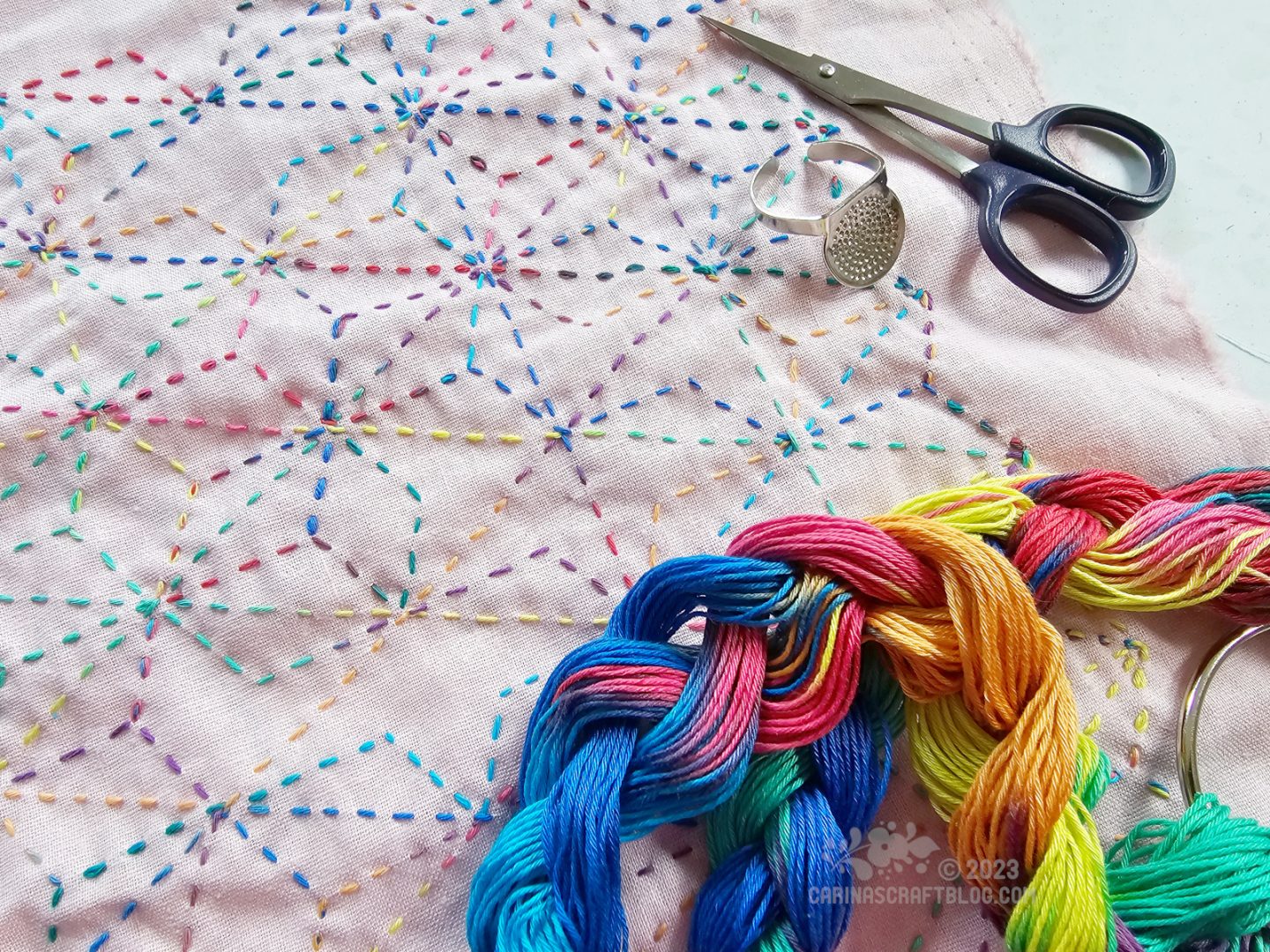 Close overhead view of a piece of pale pink fabric with runnig stitch in a rainbow coloured thread. In the foreground is a braided skein of the rainbow coloured thread.