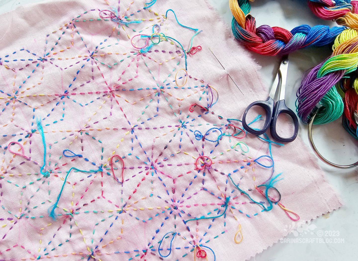 Close overhead view of a piece of pale pink fabric with running stitch in a rainbow coloured thread. In the top right corner is a braided skein of the rainbow coloured thread.