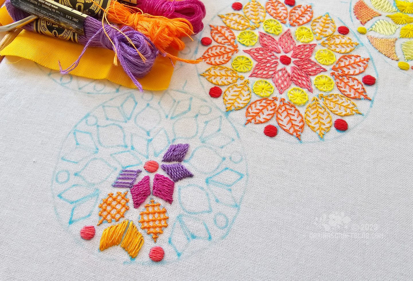 Close overhead view of a partially embroidered mandala inspired design using magenta, purple and orange colours.