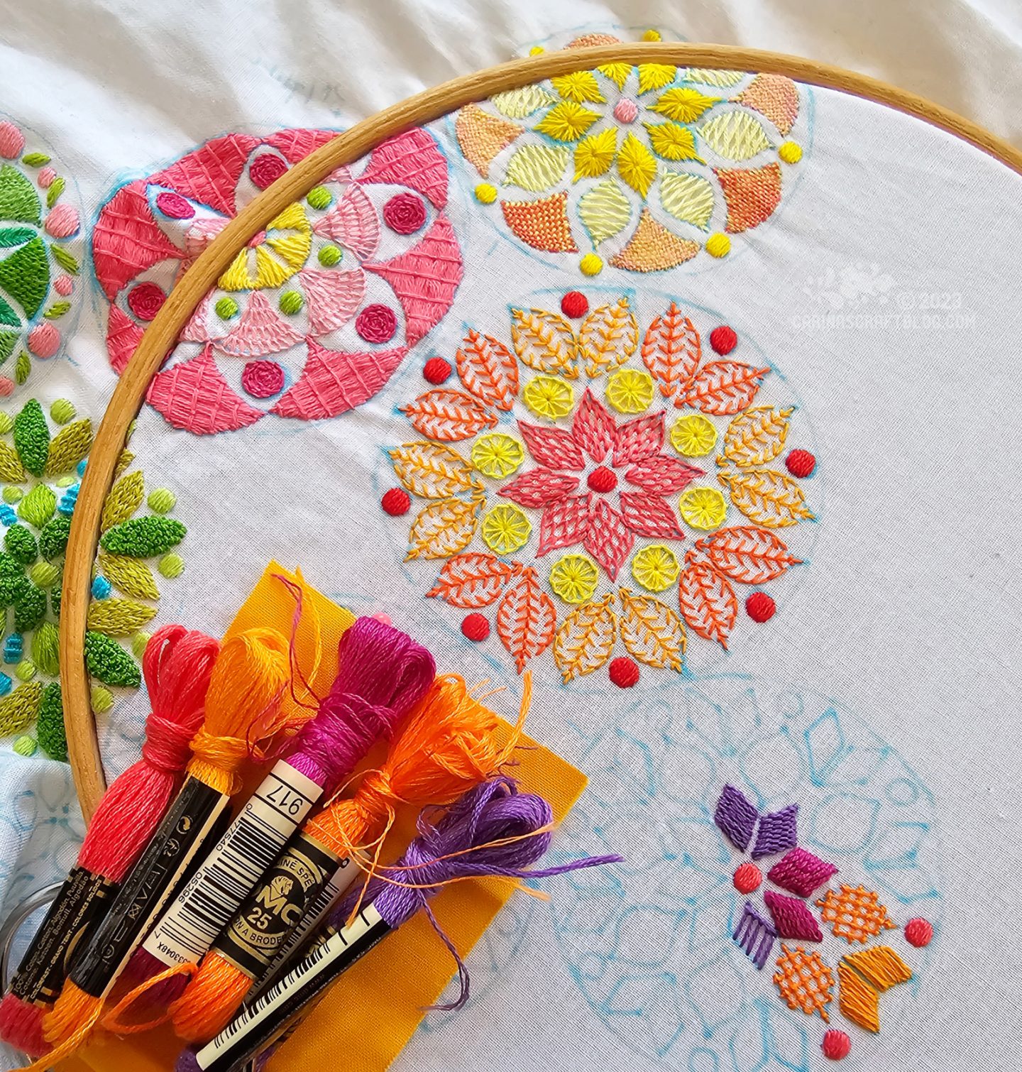 Overhead view of a large embroidery hoop with white fabric. On the fabric are embroidered several small mandala inspired designs in various colours.