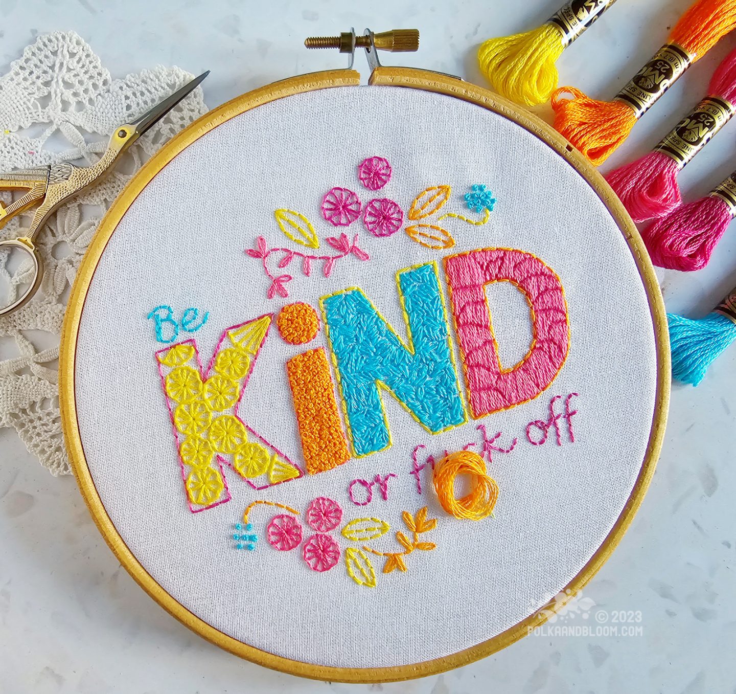 Overhead view of an embroidery hoop with white fabric. On the fabric is embroidered be kind or fuck off, using yellow, orange, pink and turquoise colours.