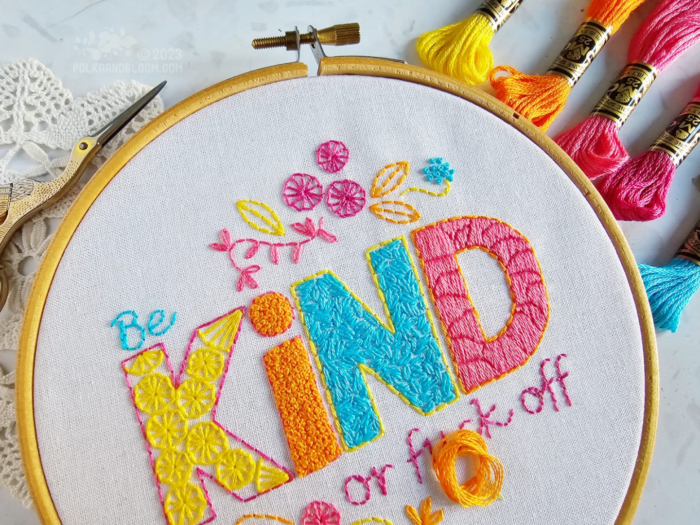 Close view of an embroidery hoop with white fabric. On the fabric is embroidered be kind or fuck off, using yellow, orange, pink and turquoise colours.