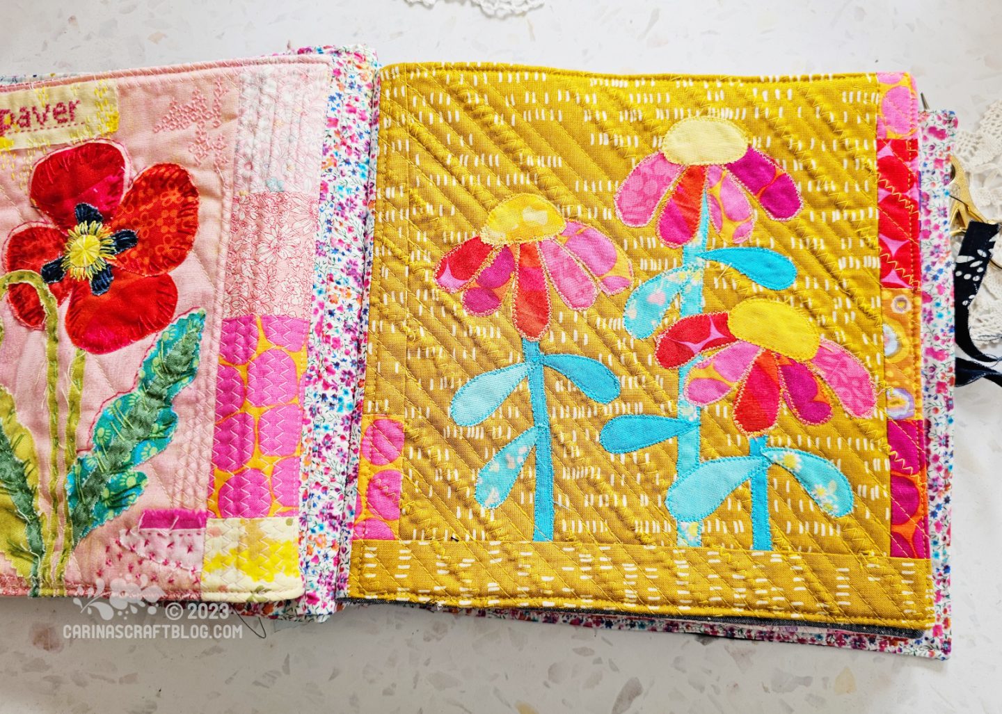 Overhead view of a spread in a textile book. Partial view of the left hand page shows a poppy on a pink background. The almost full view of the right hand page shows echinacea inspired flowers in yellow and pink with turquoise stems and leave, on a mustard yellow background.