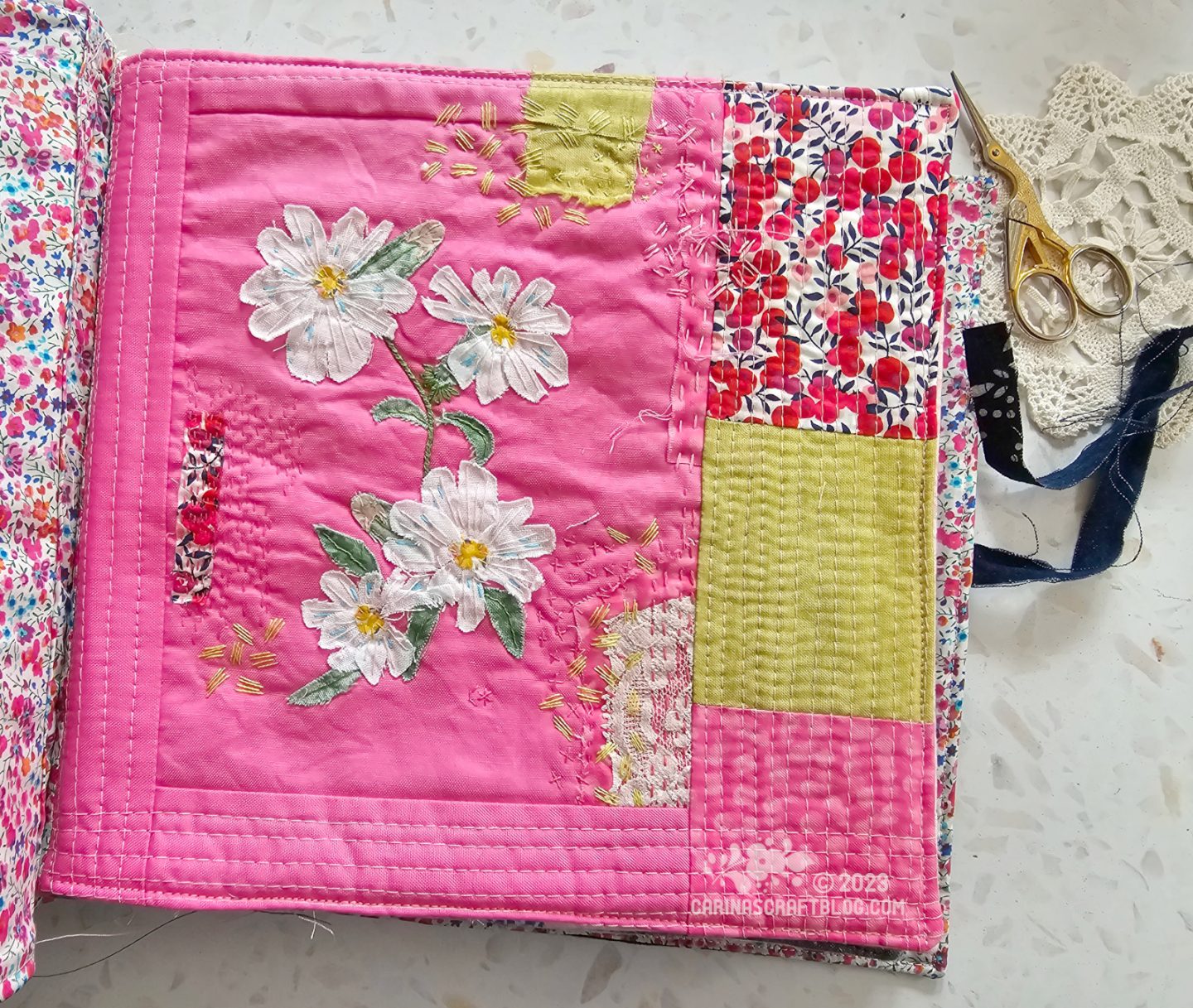 Overhead view of a page in a textile book. The page is mainly hot pink with a few patches of varying sizes, in lime green and red. Just left of the middle is an appliqué of a stem with four white flowers.