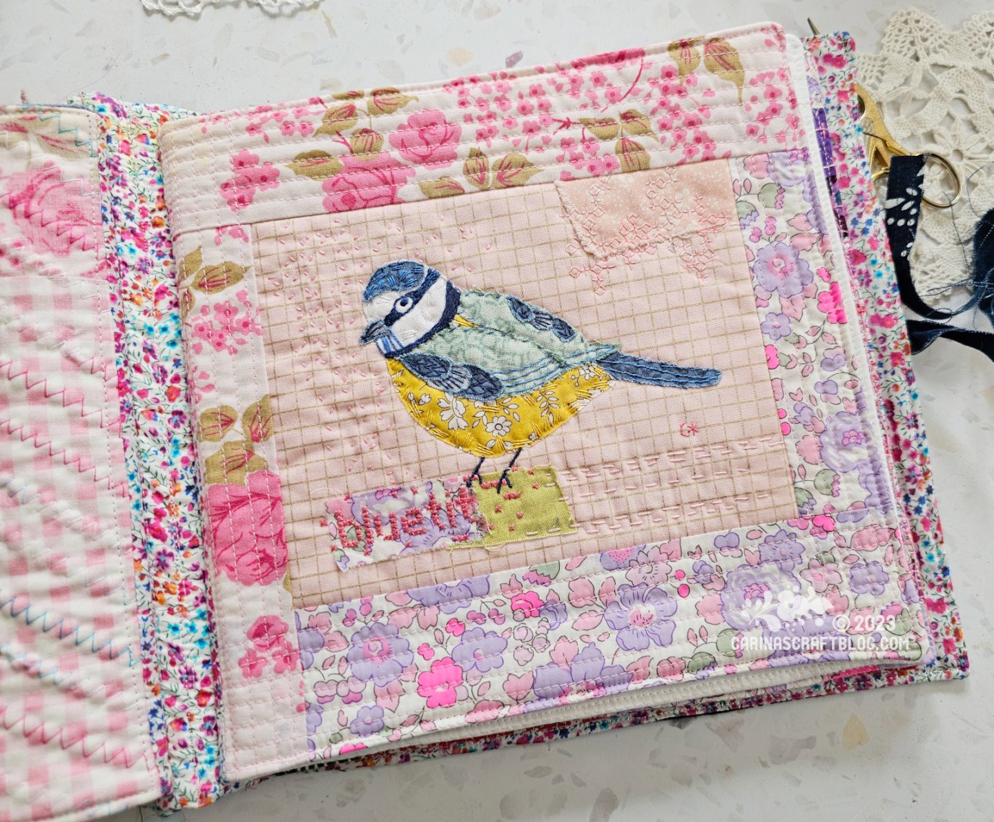Overhead view of a page in a textile book. The background is pale pinks and lilac. In the centre is an appliqué and hand embroidery blue tit.