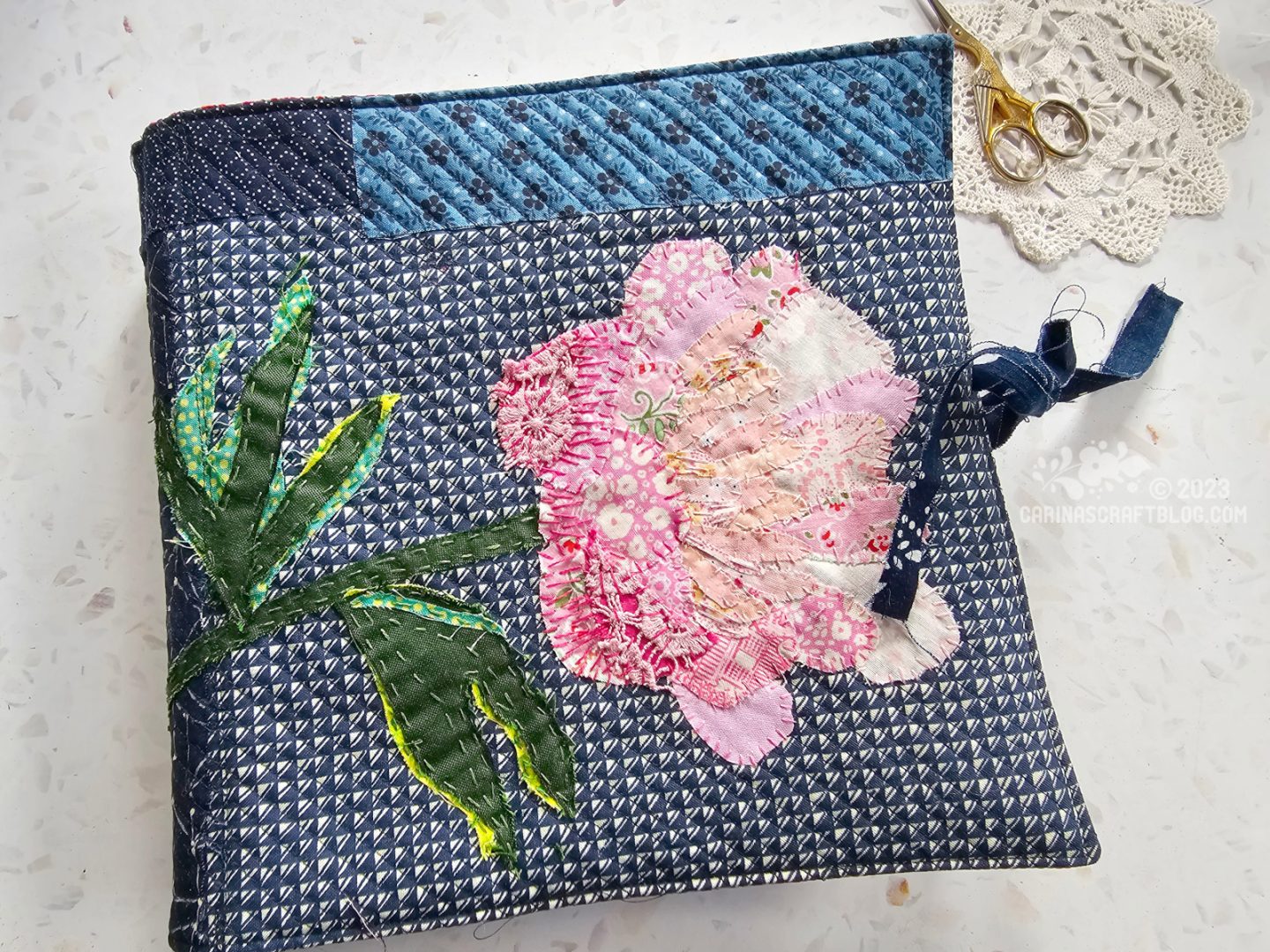 Overhead view of the front cover of a textile book. A large pink flower is appliquéd on a background of dark blue fabrics.