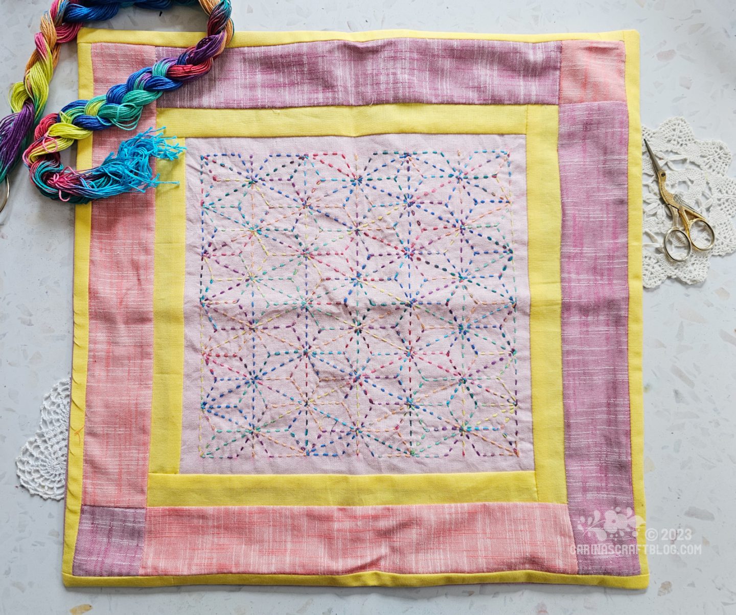 Overhead view of a cushion cover. It has an outer border in pink colours, a narrow inner border in bright yellow and a central panel in light pink with multicolour embroidery.