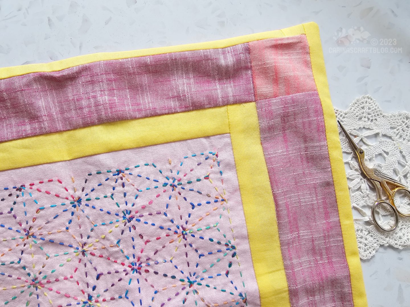 Partial, close overhead  view of a cushion cover. It has an outer border in pink colours, a narrow inner border in bright yellow and a central panel in light pink with multicolour embroidery.