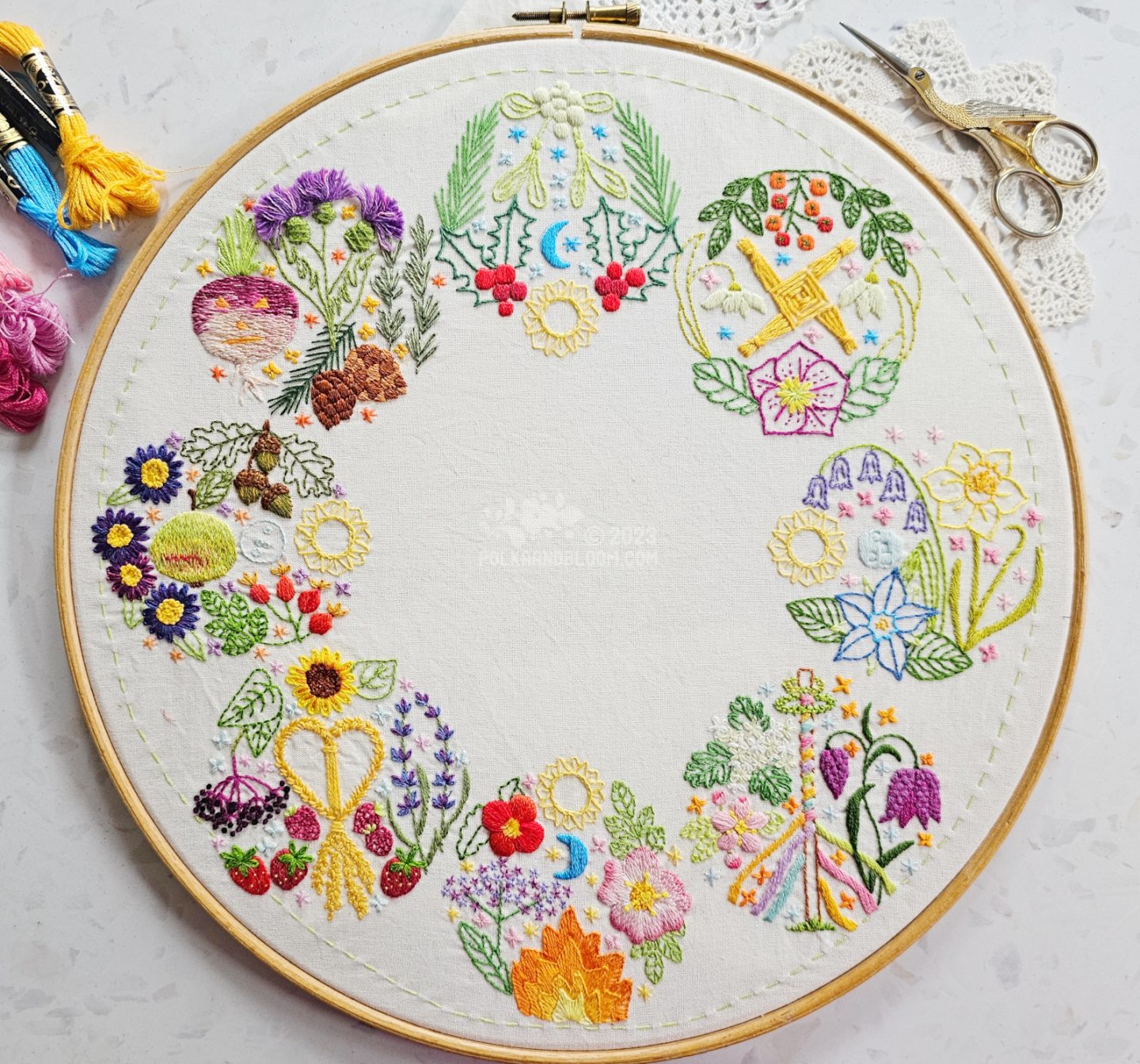 Overhead view of an embroidery in a wooden embroidery hoop. The embroidery is made up of 8 circular motifs arranged in a large circle. Each small circle depicts various elements of the 8 seasons in the Celtic Wheel of The Yea.