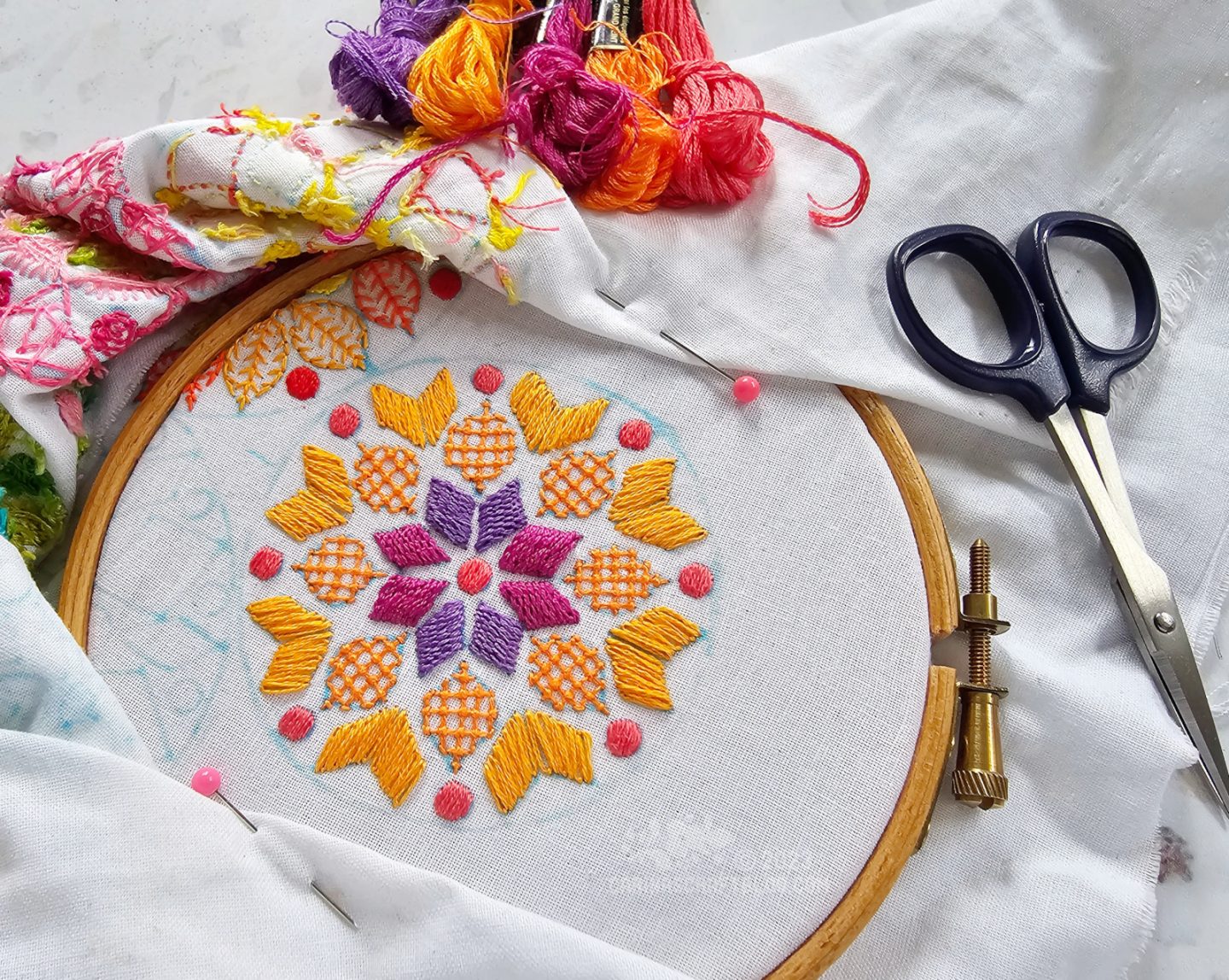 Overhead view of white fabric stretched in a wooden embroidery hoop. On the fabric is embroidered a mandala inspired design in purple and orange colours. 