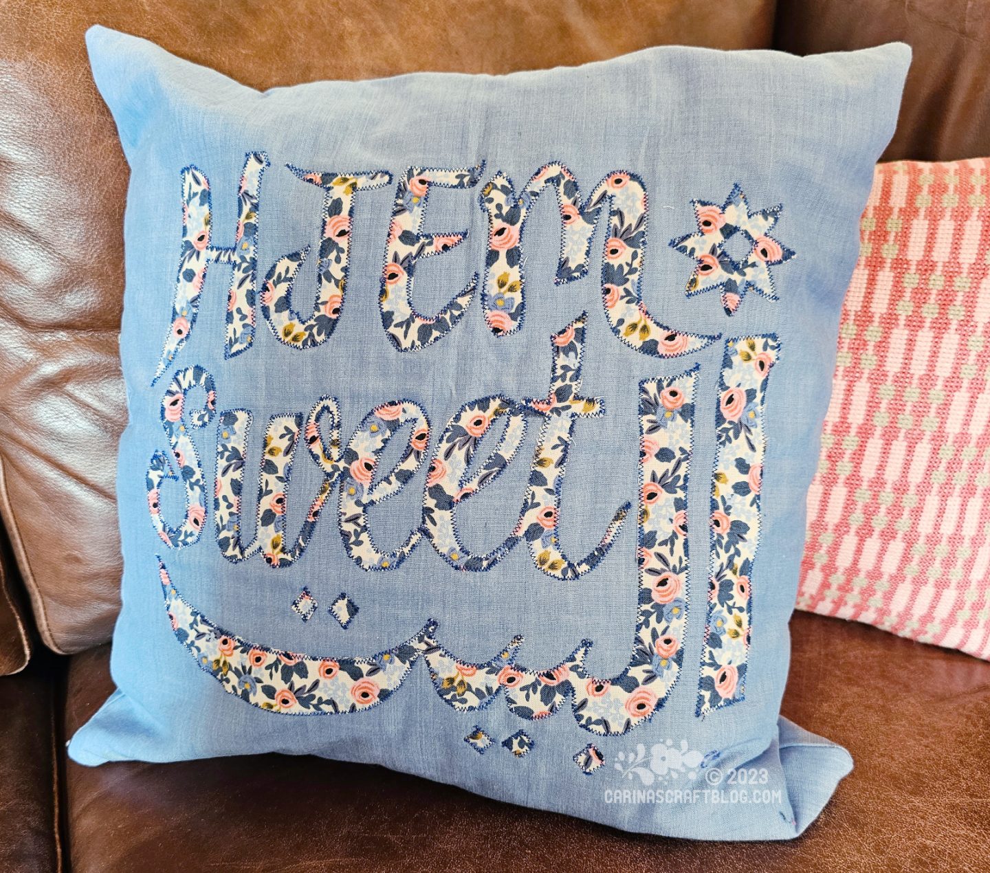 A picture of a light blue cushion with the text hjem sweet albayt appliquéd in a blue and pink floral fabric.