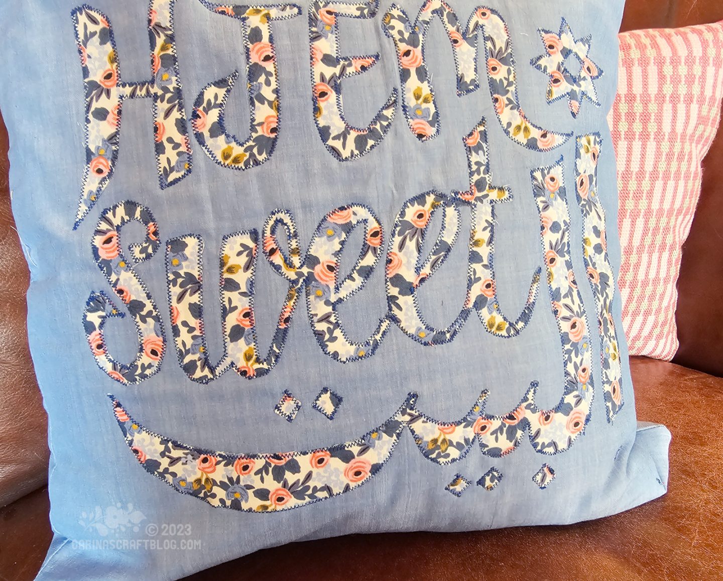 Close up view of a light blue cushion with the text hjem sweet albayt appliquéd in a blue and pink floral fabric.