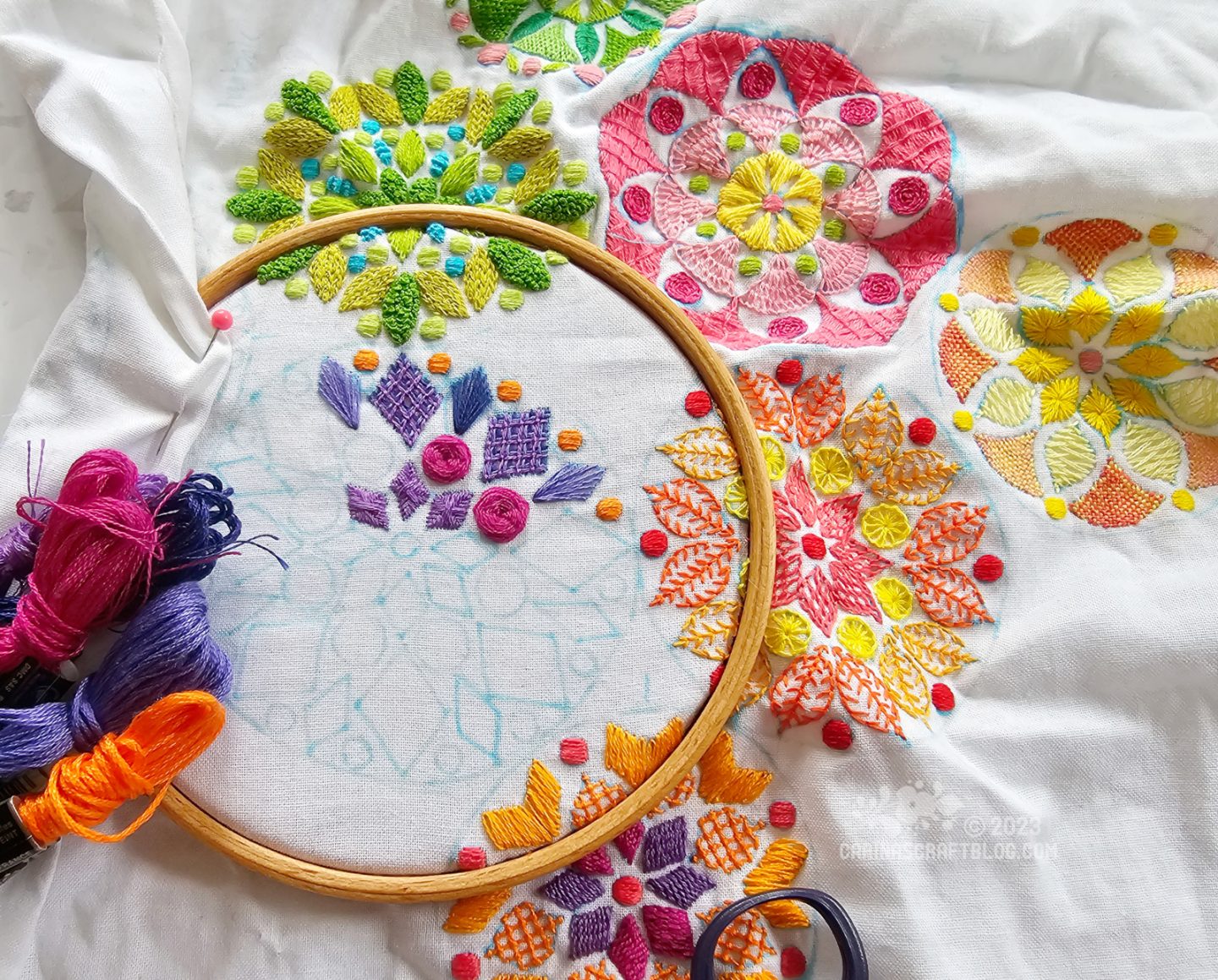 View from above of a large piece of white fabric, partially stretched in a wooden embroidery hoop. On the fabric are embroidered several mandala inspired designs in different colours, green, pink, yellow and orange.