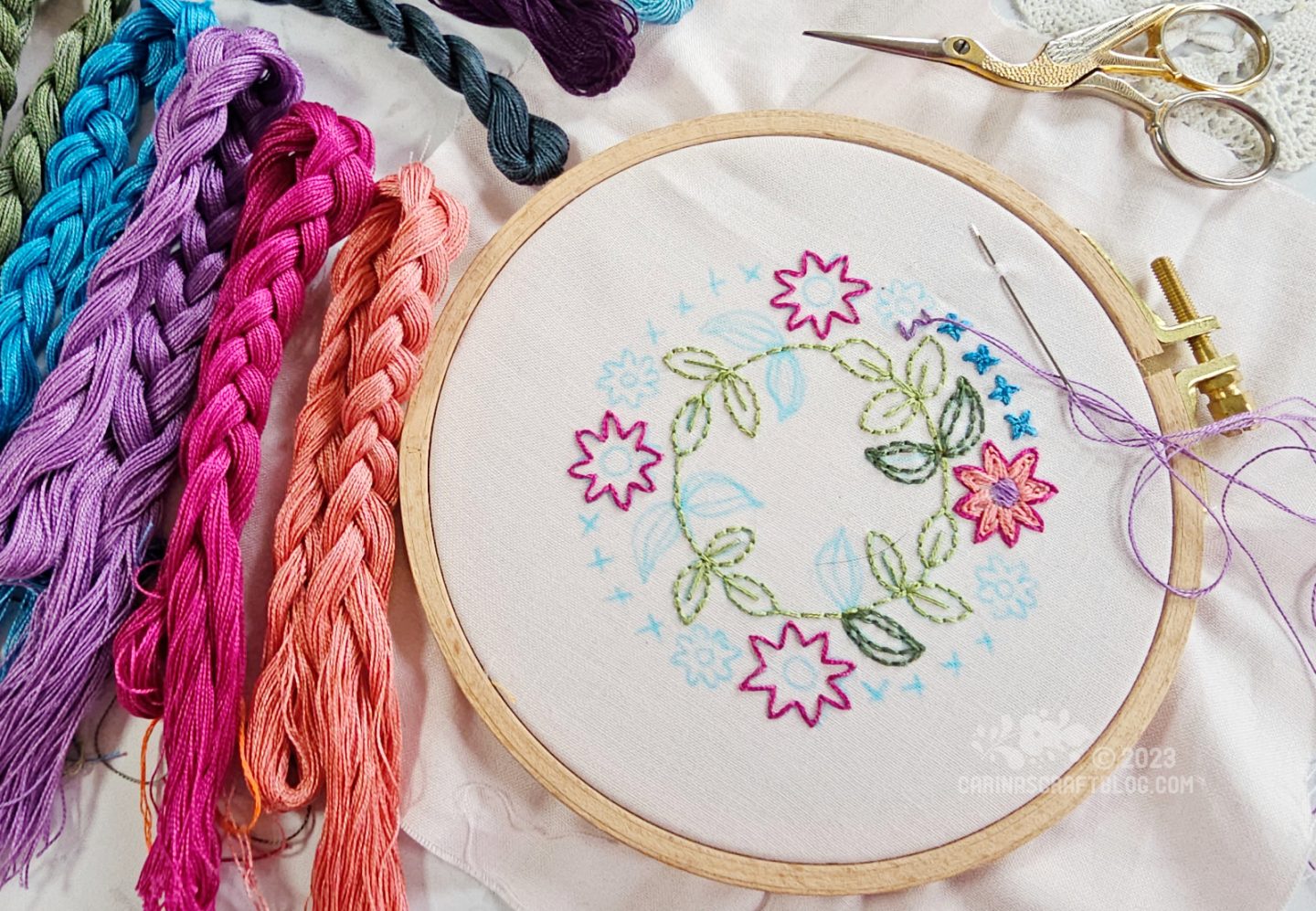 Overhead view of a small embroidery hoop with pale pink fabric. On the fabric is a partially embroidered wreath design. To the left of the hoop are several large skeins of embroidery thread in jewel colours.