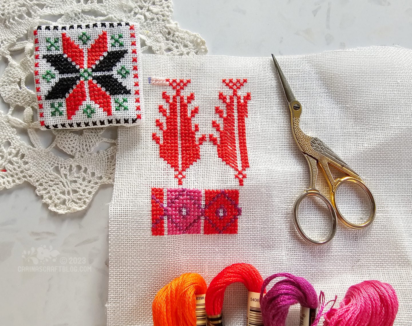 Close overhead view of white fabric partially cross stitched with red and pink colours.
In the top left corner is a small square fabric brooch made from white fabric embroidered with a star design in red and black cross stitches.