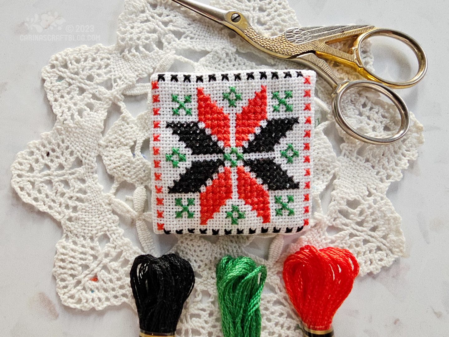 Close overhead view of a small square fabric brooch made from white fabric embroidered with a star design in red and black cross stitches.