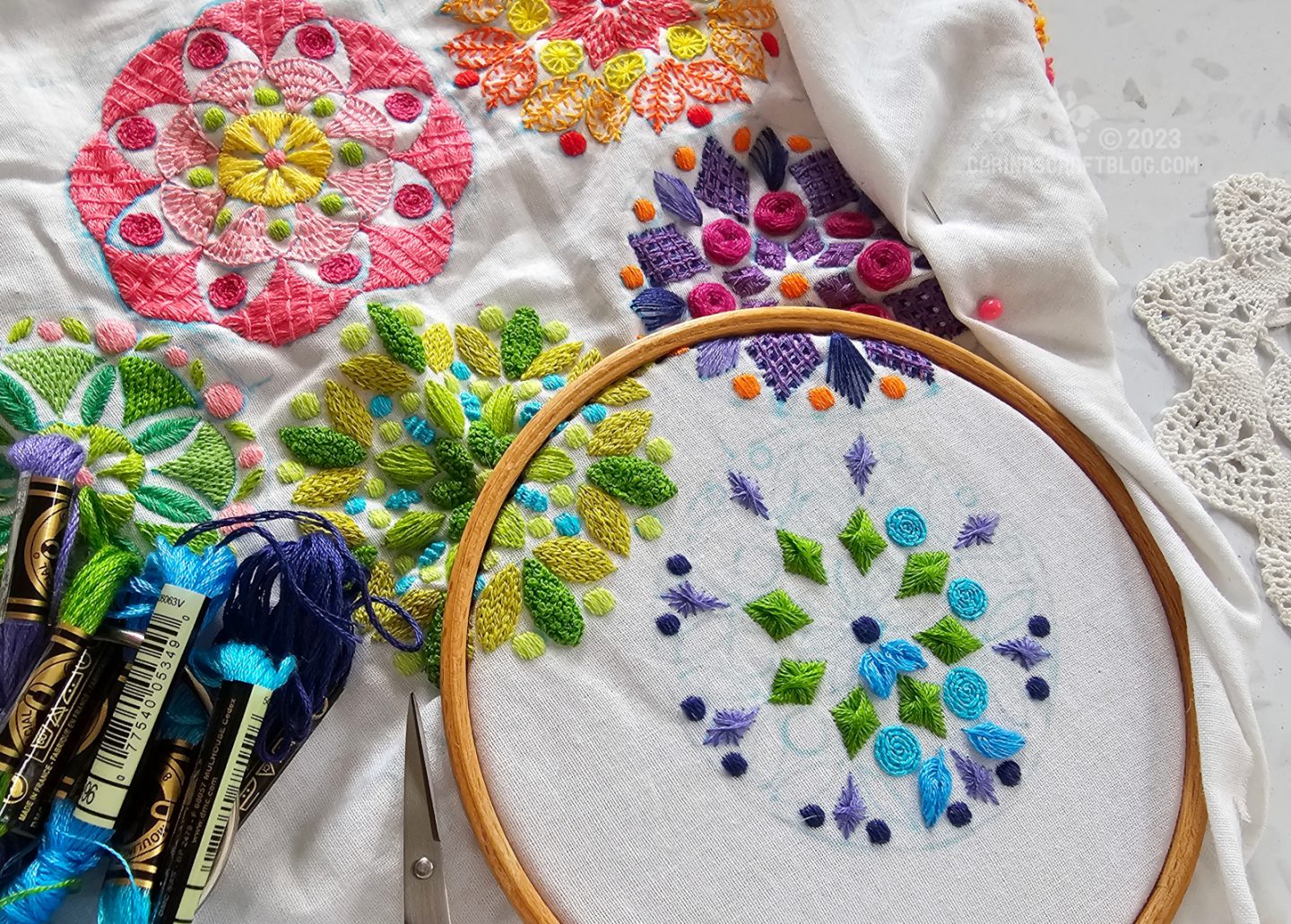 Close overhead view of white fabric part of white is stretched in a wooden hoop. In the hoop is a half finished mandala inspired motif worked in green and blue colours. Partially obscured by folds in the fabric are similar designs stitched in multiple colours.