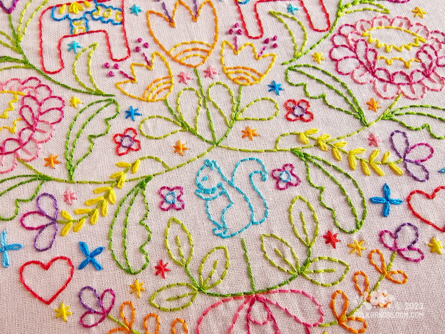 Close up view of light pink fabric stitched with a Scandi inspired design in many colours with Dala horse, flowers and leaf motifs. In the centre of the closeup is a small turquoise squirrel.