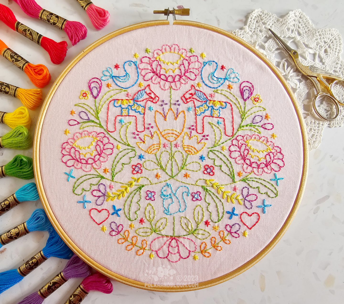 Overhead view of a round wooden embroidery hoop. In the hoop is light pink fabric stitched with a Scandi inspired design in many colours with Dala horse, flowers and leaf motifs.