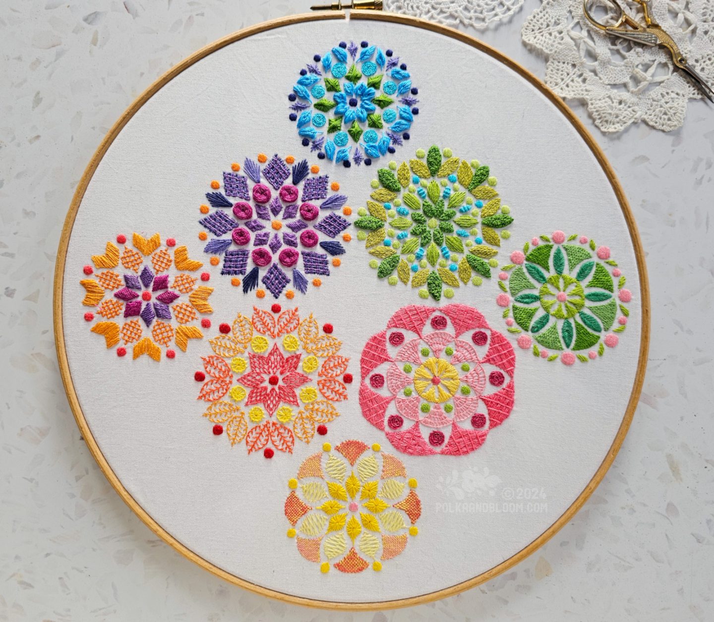 Overhead view of a large wooden embroidery hoop with white fabric. On the fabric are embroidered eight small mandala inspired designs, each in a different dominant colour: blue, lime green, spring green, pink, yellow, orange yellow, orange and purple.