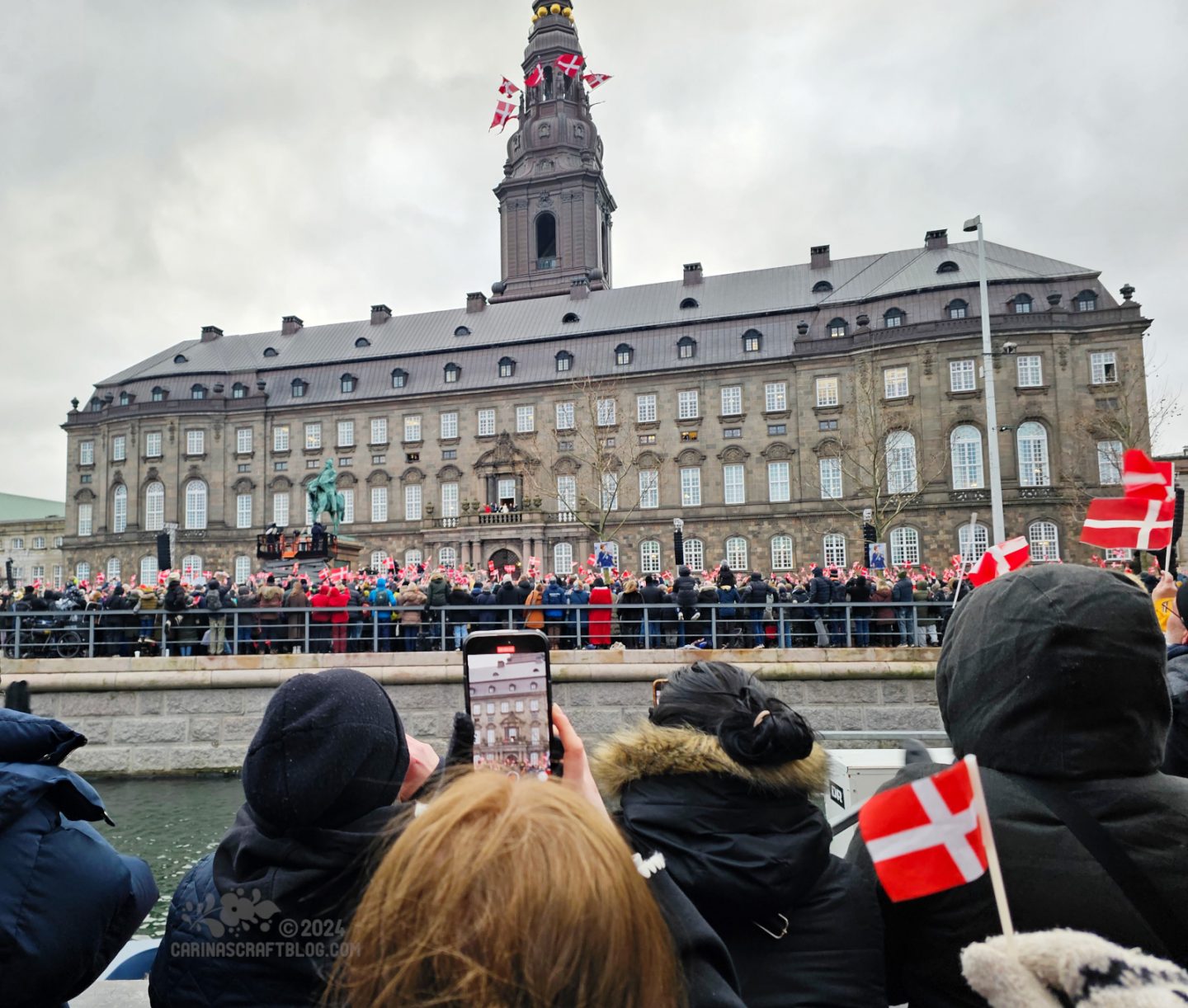 View of the Danish parliament building, Christiansborg. In front of the building, and in the foreground of the picture, are lots of people waving the red and white Danish flag.