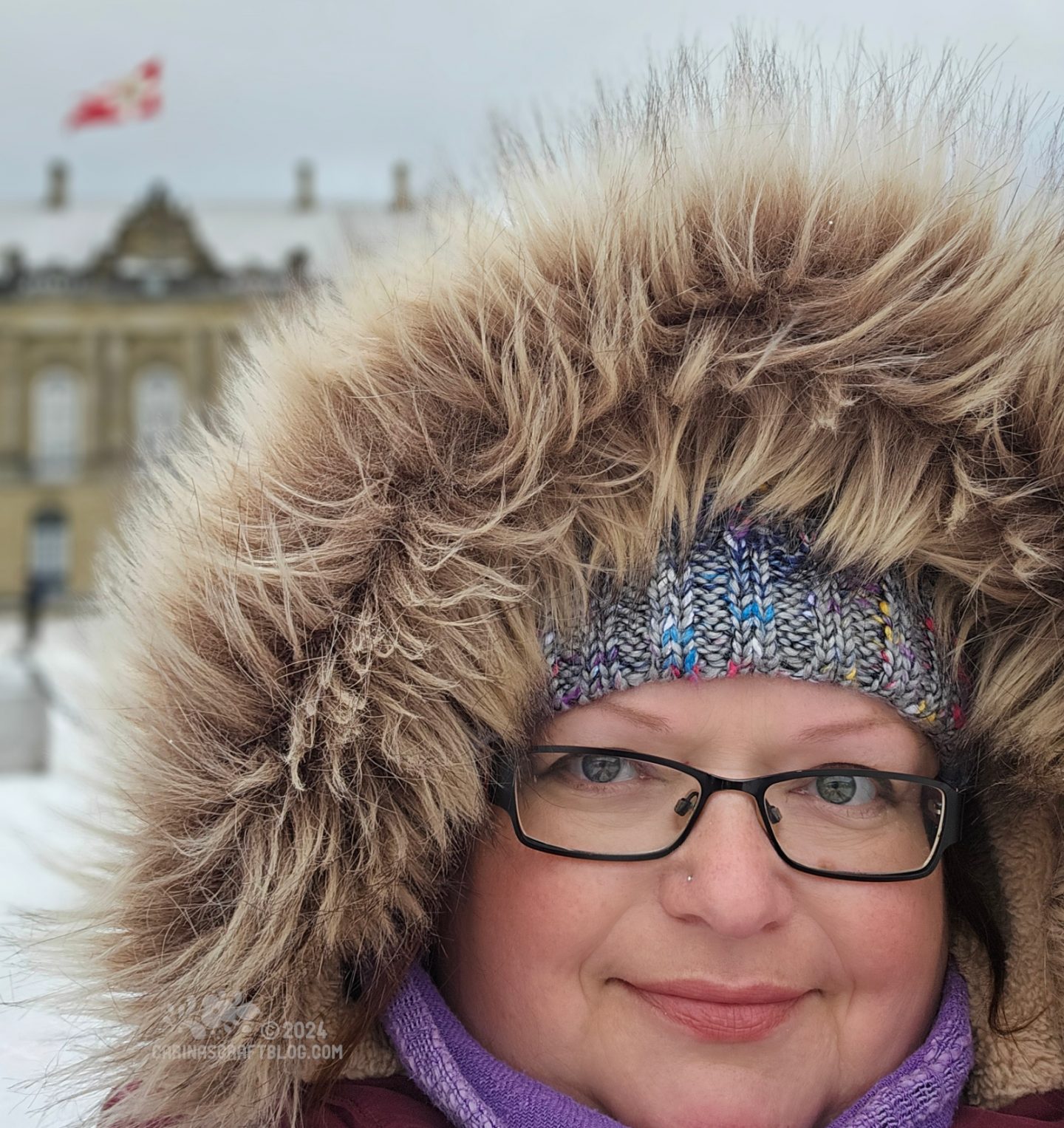 Close view of a woman's face. She is wearing glasses, a woolly hat pulled down to her eyebrows and a furry hood is framing her face. In the background there is snow on the ground and dark grey clouds.