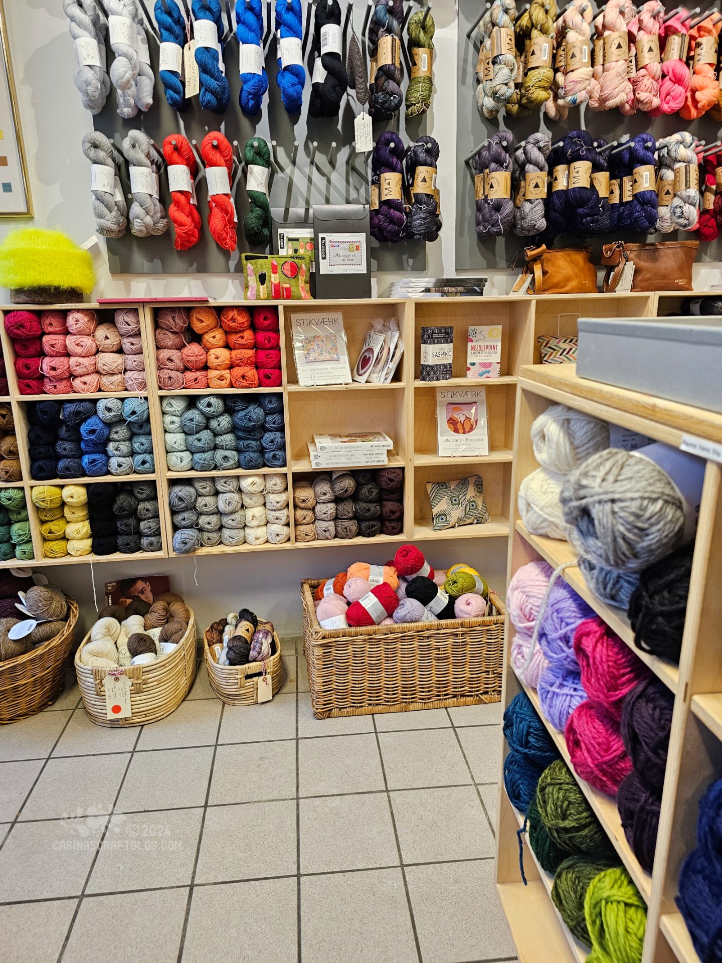 Interior view of a yarn shop. Yarn in different colours fill baskets on the floor, hangs on the wall and fill cubbies on the wall.