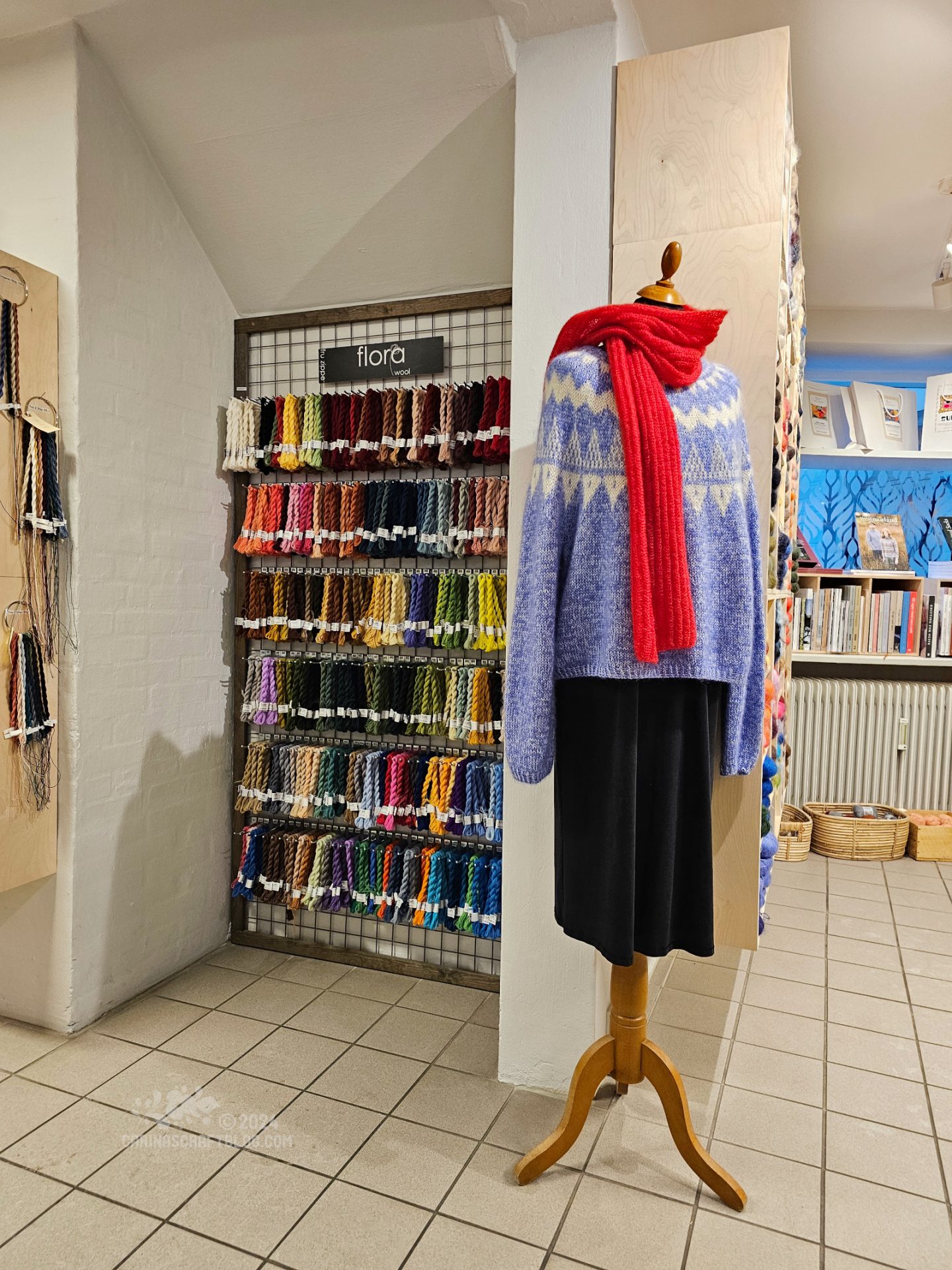 Interior view of a yarn shop. Yarn in different colours hangs on the wall. A mannequin with a blue sweater and red scarf is in the centre of the image.