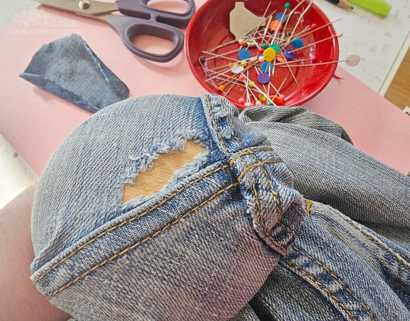 Photo of a tear in a pair of jeans. The fabric is stretched over a darning mushroom. In the background are a pair of scissors and a red bowl with pins.