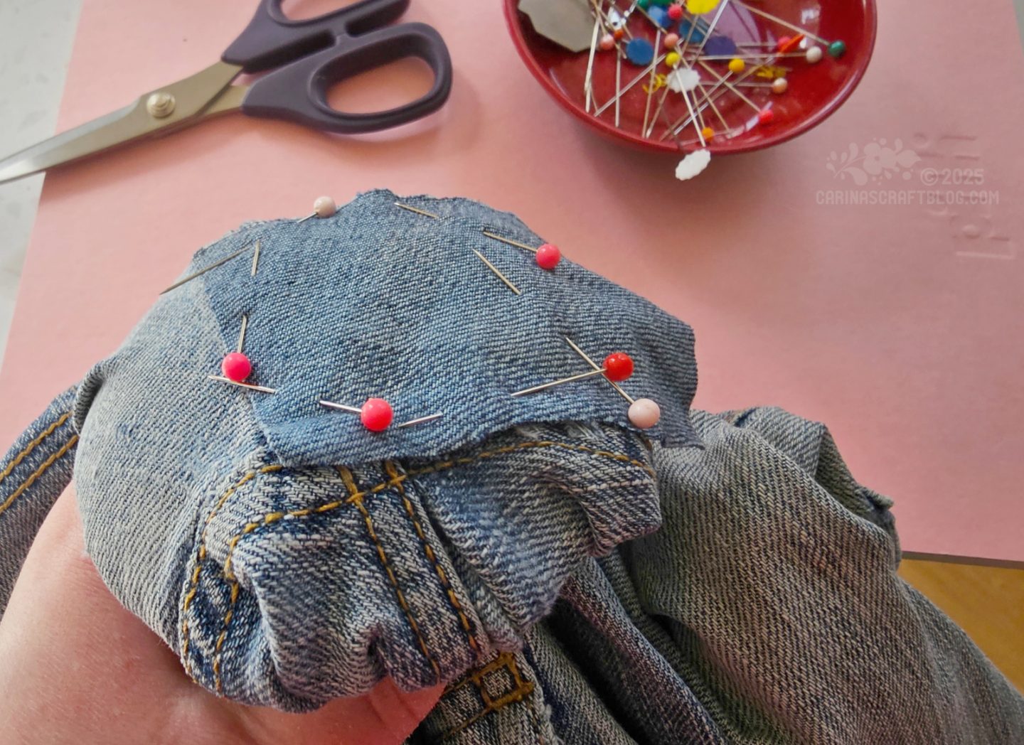 Photo of a pair of jeans. A section of the jeans is
stretched over a darning mushroom. A small patch of similar fabric is pined onto the jeans. In the background are a pair of scissors and a red bowl with pins.