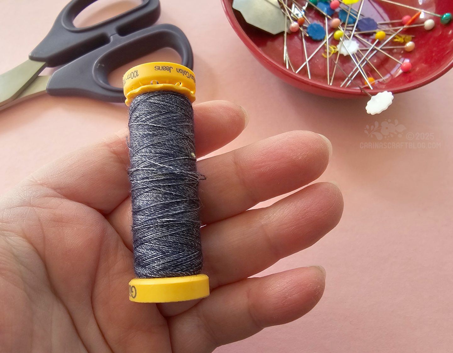 Photo of an open hand holding a variegated dark blue spool of sewing thread. In the background are a pair of scissors and a red bowl with pins.
