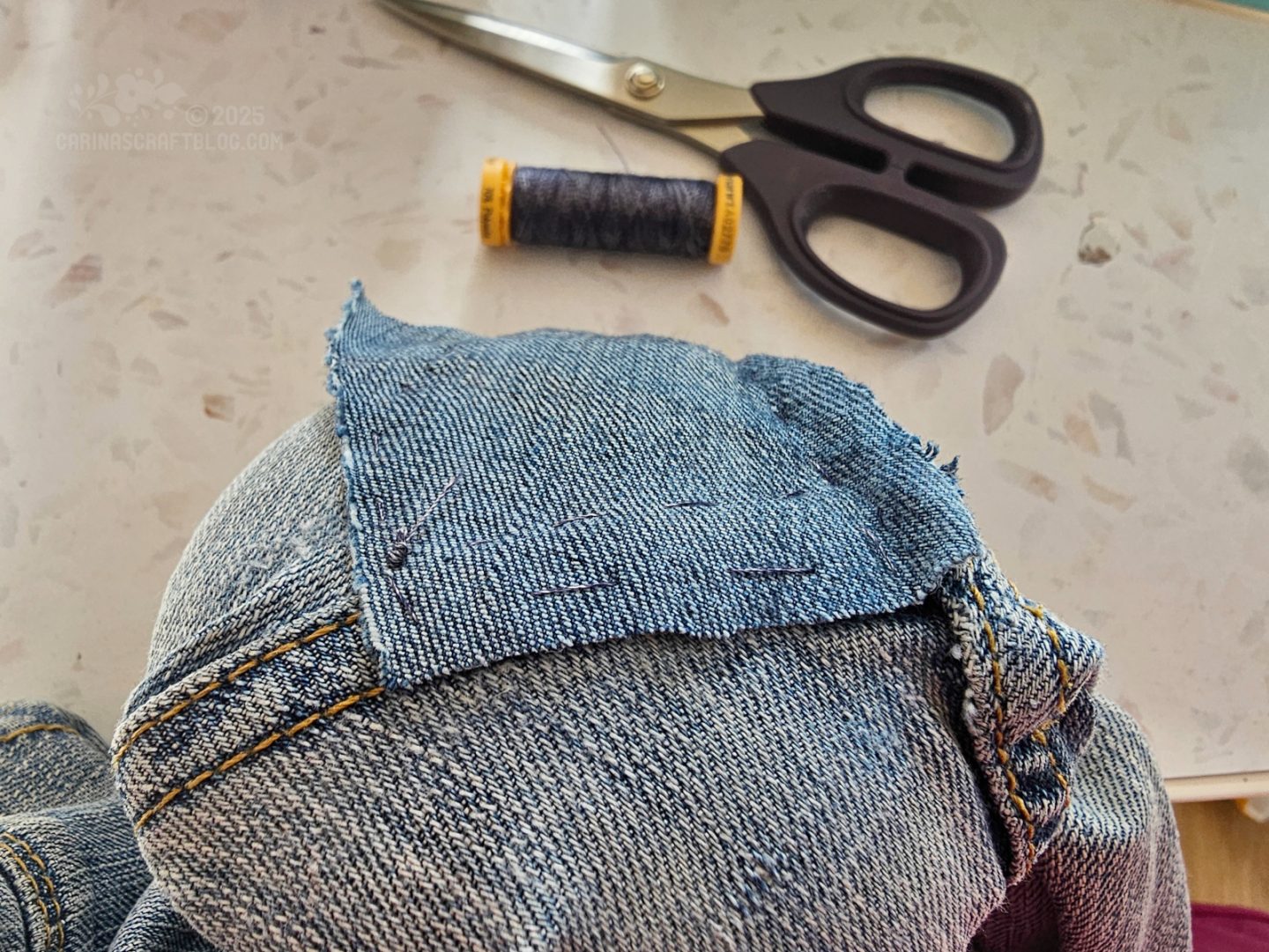 Photo of a pair of jeans. A section of the jeans is stretched over a darning mushroom. A small patch of similar fabric is stitched onto the jeans. In the background are a pair of scissors and a spool of dark blue thread.