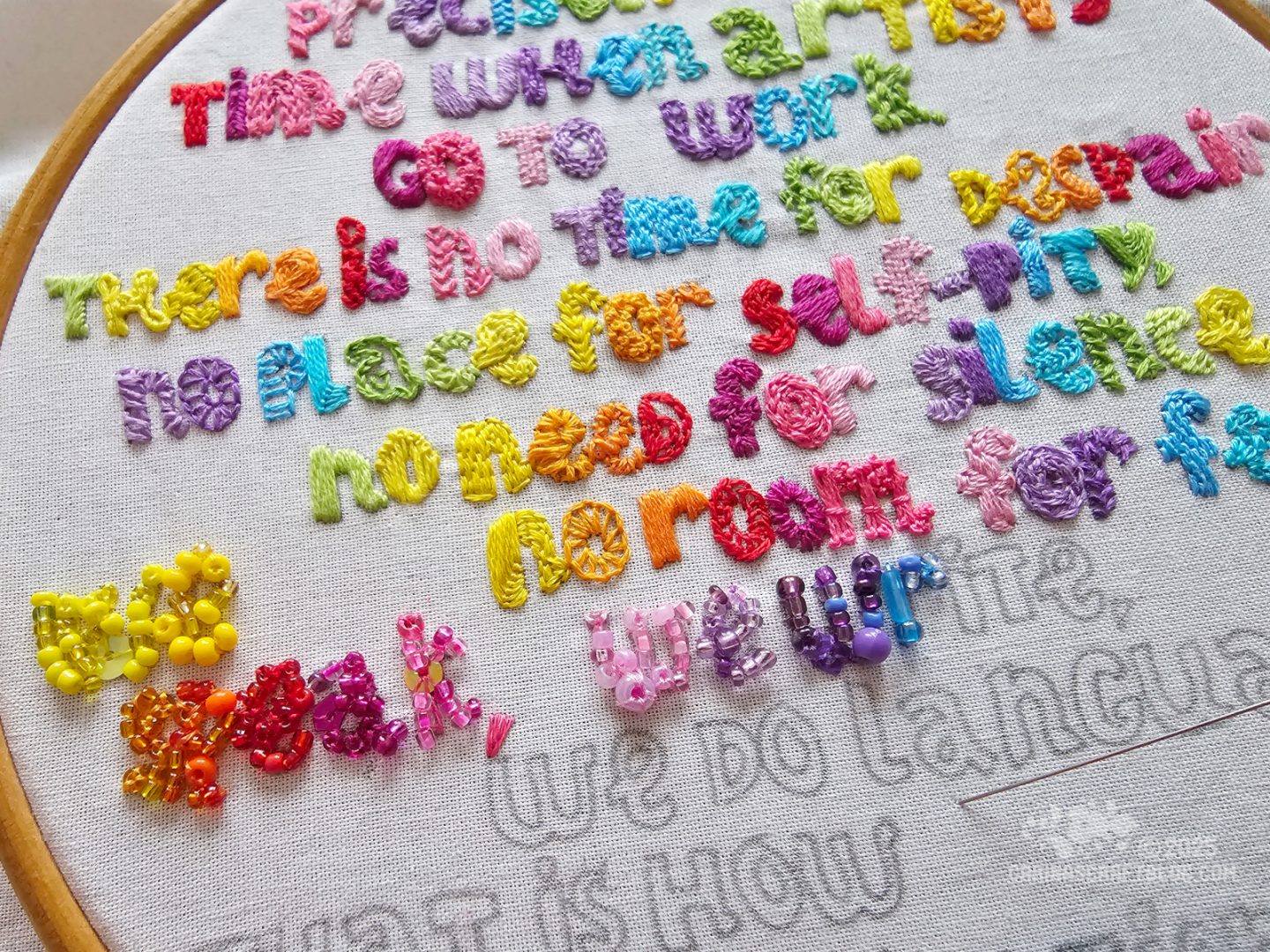 Photo, close view of white fabric stretched in a wooden hoop. On the fabric is a quote, partially stitched with lots of bright colours. Ten of the letters have been filled with beads.