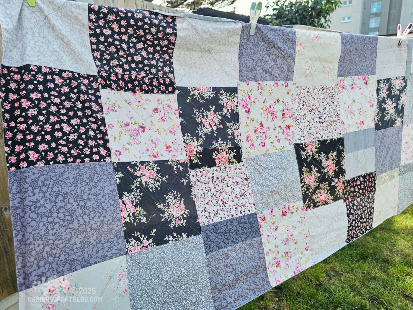 Photo of a blanket hanging on a washing line in a garden. The photo is cropped very close to the blanket. The blanket is made of fabric squares in different patterns and shades of black and grey.