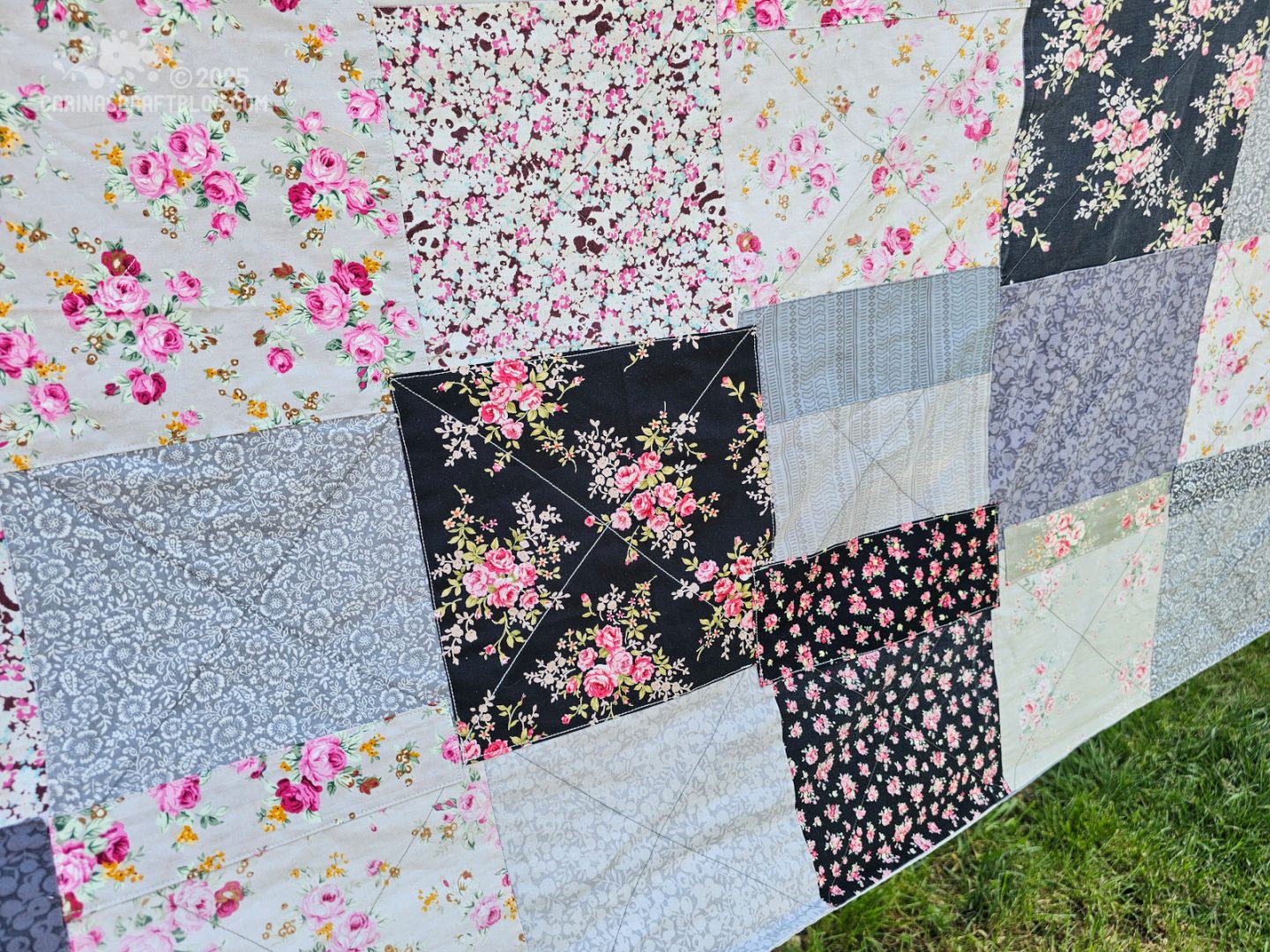 Close up photo of a blanket hanging on a washing line in a garden. The photo is cropped very close to the blanket. The blanket is made of fabric squares in different patterns and shades of black and grey.
